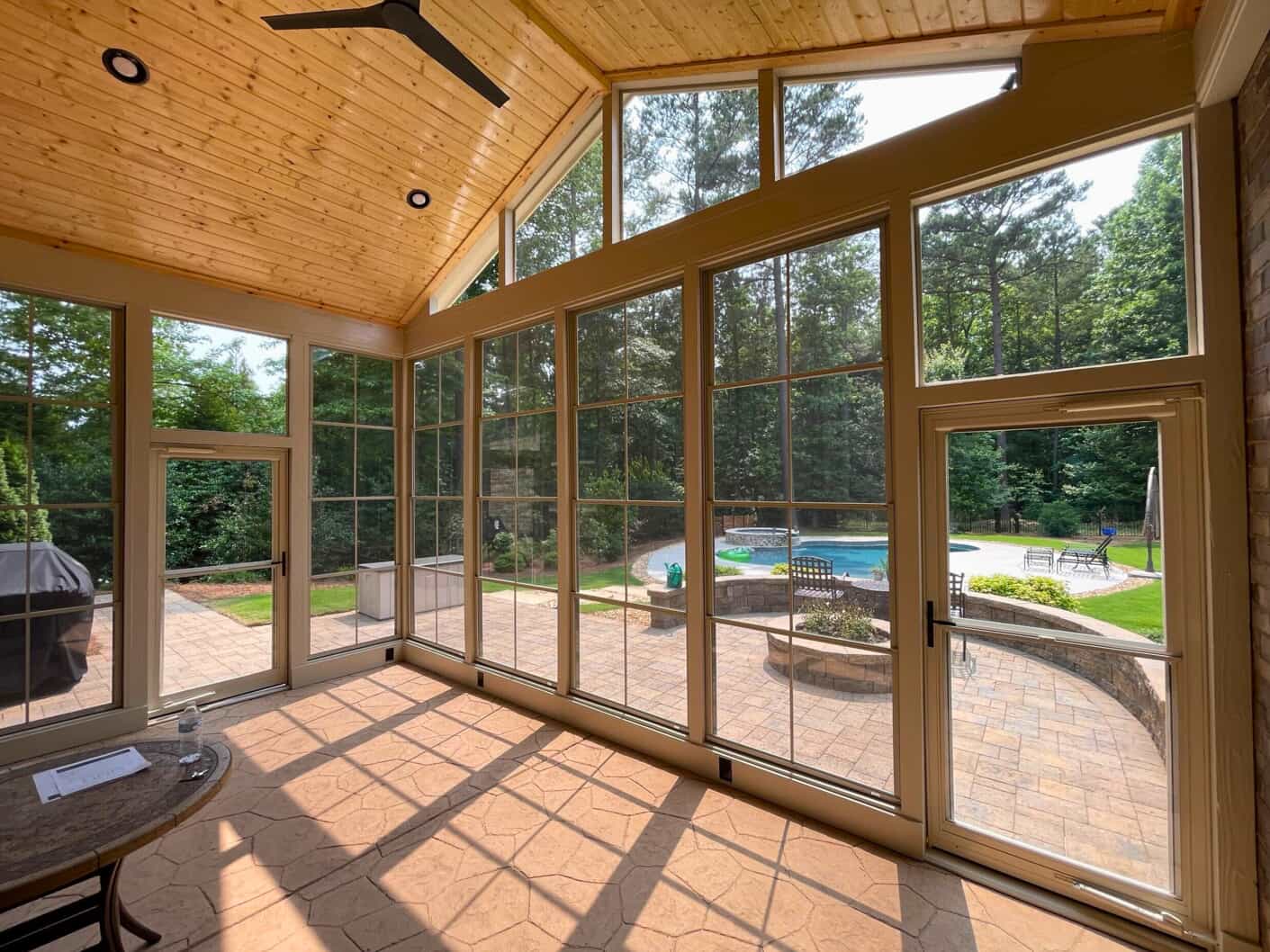 A sunroom with light-colored tile flooring, large windows, and a wood plank vaulted ceiling with a black ceiling fan. The windows look out onto a paved patio with a fire pit and a swimming pool in a lush green backyard.